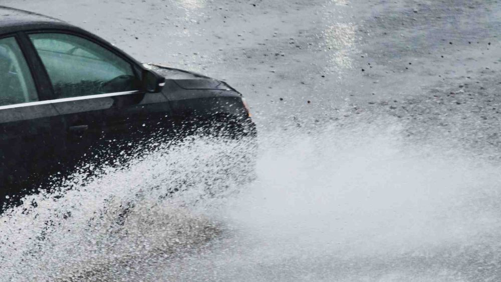 Heavy rain, rising sea levels, car driving through deep standing water on a wet road, creating a large white splash that partially obscures the vehicle.