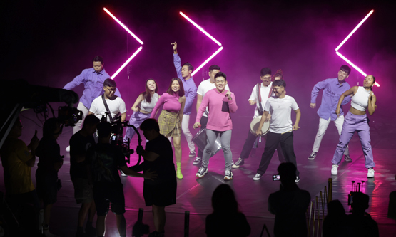 Live show production featuring a large group of performers on a stage illuminated by vibrant pink neon lights.